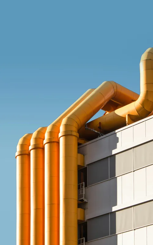 Mehrere orange Industrierohre verlaufen an der Fassade eines Hochhauses vor blauem Himmel